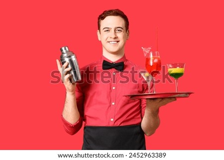 Young male bartender with shaker and glasses of cocktail on red background Royalty-Free Stock Photo #2452963389