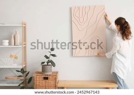 Young pretty woman hanging picture on white wall in room