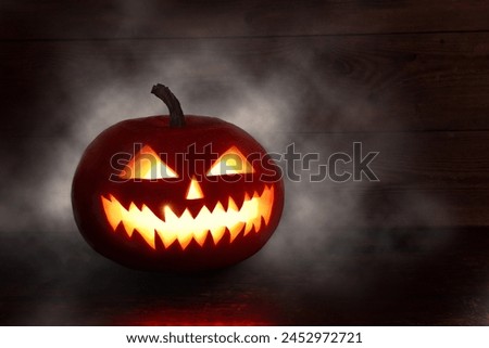 Scary Halloween pumpkin with eyes glowing inside at dark background. Spooky Halloween pumpkin, Jack O Lantern, with an evil face and eyes on a wooden table with a misty gray background. 