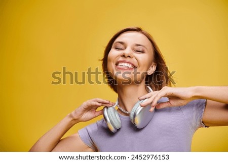 debonair joyous woman with short blonde hair and headphones enjoying music on yellow backdrop Royalty-Free Stock Photo #2452976153