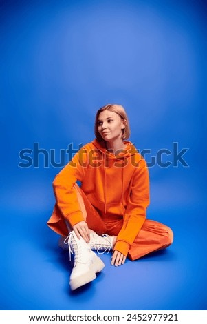 debonair cheerful woman with short hair in vibrant orange hoodie posing actively on blue backdrop Royalty-Free Stock Photo #2452977921
