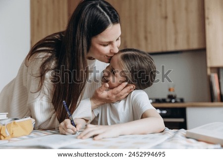 A mother helps her young daughter with homework, both focused on a workbook at a table.