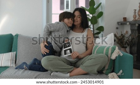 Happy Mother and child posing with ultrasound picture of unborn baby brother during third trimester of pregnancy seated at home couch. Mom and son smiling at camera