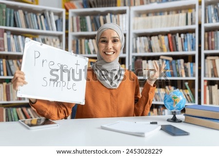 Cheerful Muslim woman teaches English grammar, using a whiteboard in a library setting. Her engaging smile and clear presentation enhance the educational atmosphere.