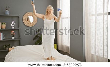 A happy caucasian woman dances on a bed in her bedroom, holding a blue credit card and a smartphone, conveying joy and leisure indoors.