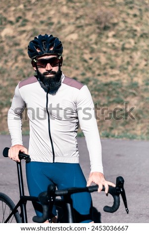 A male cyclist standing next to a bicycle, wearing a helmet and white and brown jacket. Looking at the camera.