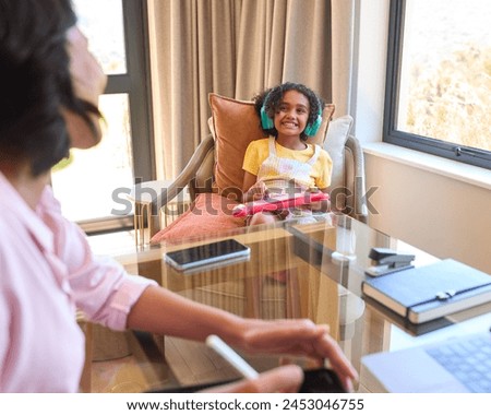 Mother Working From Home On Digital Tablet As Daughter Listens To Music And Plays In Background