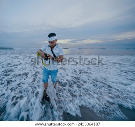 Full length of asian man landscape photographer wearing a udeng or white headband with camera in his hands looks at photos on the camera display with a beach background at sunset.