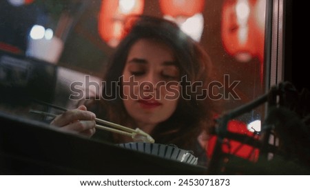 Reflective woman enjoys Chinese meal with chopsticks observing night city street through window. Appealing lady takes well-deserved break after workday in Asian restaurant during late evening Royalty-Free Stock Photo #2453071873