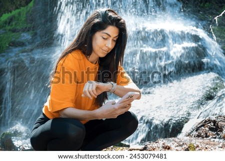 Fit Woman Looking her Smartwatch in the summer mountain on a waterfall background 