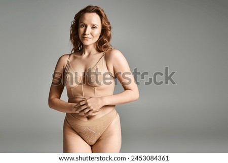 A striking woman with natural beauty poses gracefully in a tan bodysuit, exuding elegance and confidence.