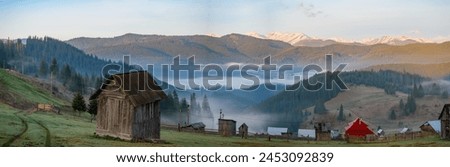 View of mountain village surrounded by peaks of Carpatian mountains with wooden cabins and faint trail of an automobile tires on dews grass. Mist gently blankets mountain valley village in early hours Royalty-Free Stock Photo #2453092839