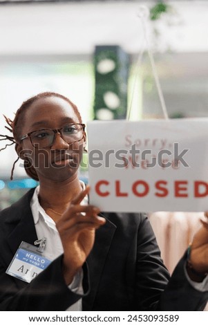 Woman holding closed sign on fashion clothes store front door, informing customers about business hours end. Apparel shop african american assistant hanging sign on widow