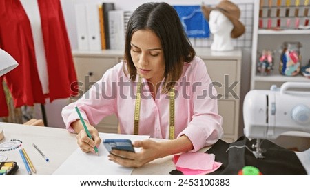 A young woman fashion designer sketching while consulting her phone in a sewing workshop.