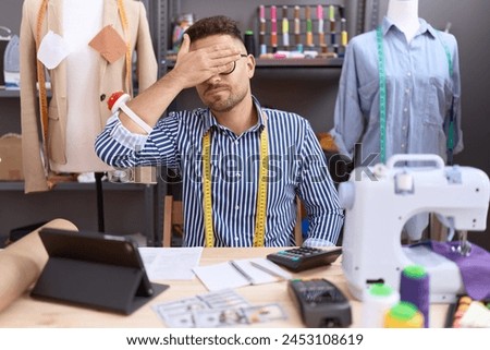 Hispanic man with beard dressmaker designer working at atelier covering eyes with hand, looking serious and sad. sightless, hiding and rejection concept  Royalty-Free Stock Photo #2453108619