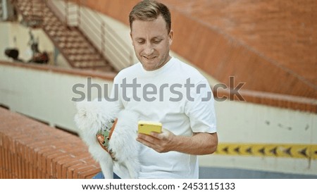 Young, confident caucasian man with his happy pet dog, smiling, sitting on a bench in the city; engrossed in texting on his smartphone, all joyful enjoying the fun of the urban outdoors.