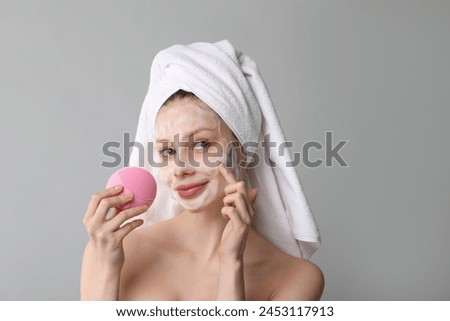 Young woman washing face with brush and cleansing foam on grey background