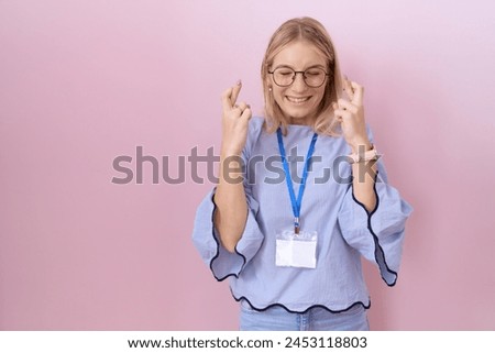 Young caucasian business woman wearing id card gesturing finger crossed smiling with hope and eyes closed. luck and superstitious concept. 
