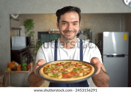 Cheerful male chef extends his arm to present a freshly baked pizza tray and graciously smiles at the camera positioned within the confines of the bustling kitchen. Royalty-Free Stock Photo #2453156291