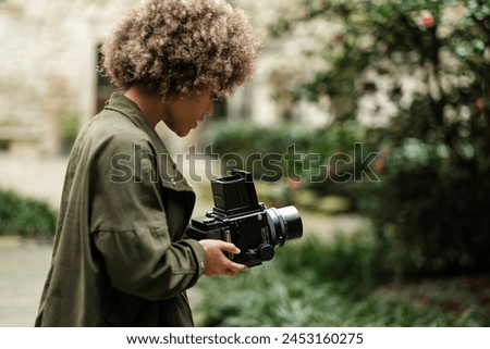 Attractive curly woman using an analog medium format camera to taking a picture in a park. She is wearing a casual dark green jacket. Traditional hobbies concept.