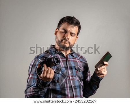 Young man indecisive between a camera and the camera of his mobile phone. Man deciding whether to use a professional camera or a mobile camera. Comparing cameras Royalty-Free Stock Photo #2453202169