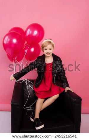 A child in a vibrant red dress and black leather jacket sits on a dark couch,surrounded by shiny pink balloons against a pink background.Concept for birthday parties, celebrations or fashion for kids.