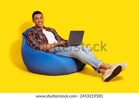 Full length photo of smart guy wear checkered shirt sitting on bean bag typing on laptop at work isolated on vibrant color background