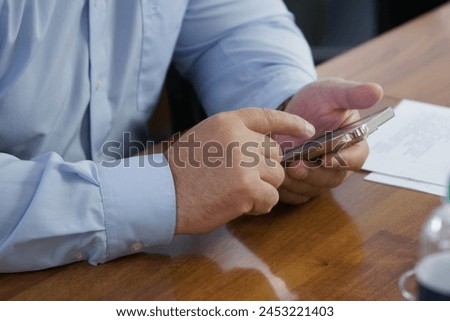 An adult male businessman in a light blue shirt is using a cell phone - smartphone while sitting at his desk. Texting or chatting. Photo. Selective focus. No face