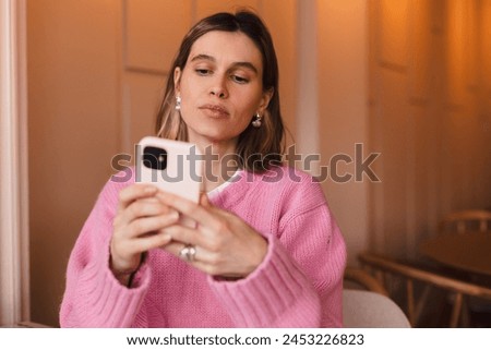 Young blonde woman using phone sits in cafe at table hold smartphone, answering texts, phone call, letters, posting photos, chatting or reading sms. Girl messaging, paying using phone.