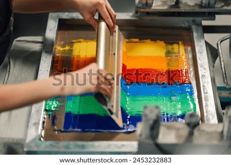 Close up of graphic technician's hands silkscreen printing with squeegee at printing shop. Cropped picture of printing shop worker screen printing and pressing ink into silk with rubber blade at shop.