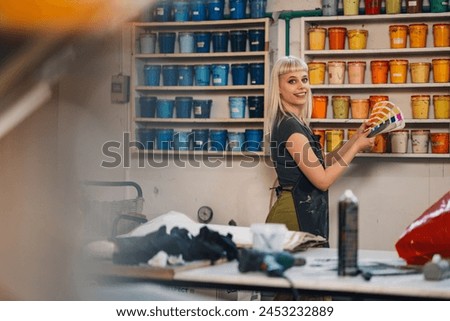 Portrait of a smiling female graphic technician standing at printing shop near shelves with ink and holding color swatch in hands while choosing paint buckets. Print shop worker with color swatch.