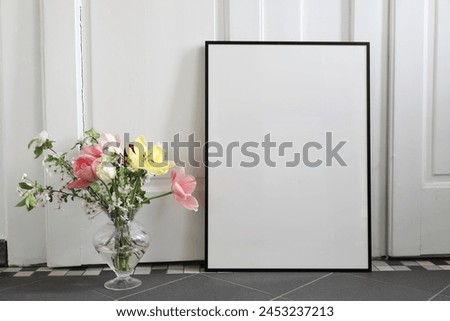 Spring still life scene. Tulips, cherry tree blossoms bouquet in glass vase. Blank black picture frame, poster mockup on grey tile floor. White wall background . Artisitic Easter decor. Interior home.