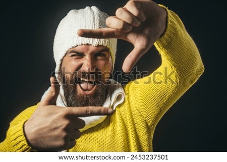 Handsome man creating frame with fingers. Bearded man using hand framing to create viewfinder. Imaginary selfie photo. Happy guy in yellow sweater, white hat and scarf makes camera frame with fingers.