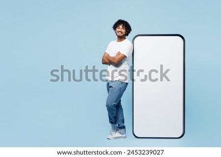 Full body side view young Indian man he wear white t-shirt casual clothes stand near big huge blank screen mobile cell phone smartphone with area isolated on plain blue background. Lifestyle concept