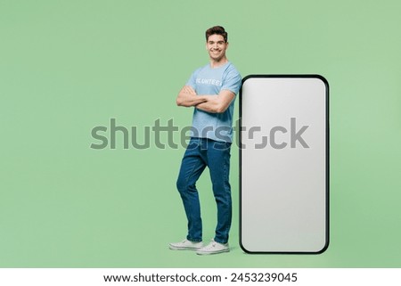 Full body happy young man wear blue t-shirt title volunteer stand near big huge blank screen area mobile cell phone isolated on plain green background. Voluntary free work help charity grace concept