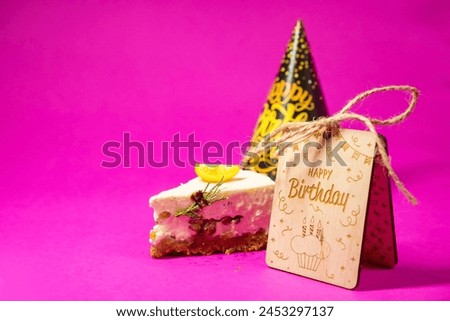 The image shows a slice of cake with a single lemon wedge on top, placed on a pink background. Next to the cake, there is a small, decorative birthday cake with a yellow frosting design and a black pa