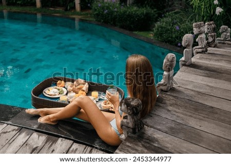 Back view of young pretty woman having Floating Breakfast tray in luxury pool hotel, young female with hat enjoy in tropical resort. Relaxing, Exotic summer travel, holiday, vacation and weekend