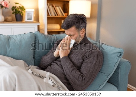 Sick sad man sits on couch at home suffers from runny nose flu disease coronavirus pandemic covid epidemic sneezes. Unwell guy feeling bad fever virus illness symptoms indoor Royalty-Free Stock Photo #2453350159