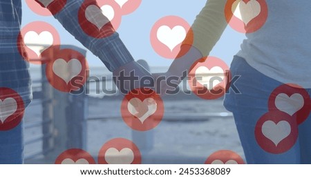 Image of red heart icons floating over mid section of couple in love holding hands by seaside. Happy Valentines Day celebration concept digitally generated image.