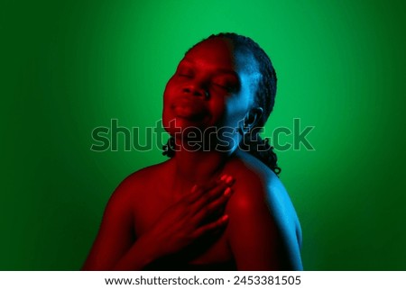 Elegant African woman with bare shoulders, well-kept spotless skin, posing on green studio background in neon light. Concept of natural beauty, ethnicity, self-care, wellness, positive emotions Royalty-Free Stock Photo #2453381505