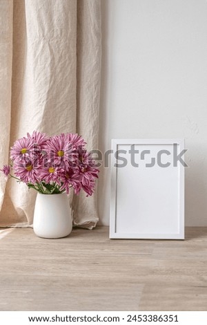 Poster mockup with empty copy space, white picture frame and pink chrysanthemum flowers on neutral beige wooden table, linen textile curtain, white concrete wall background with soft natural light.