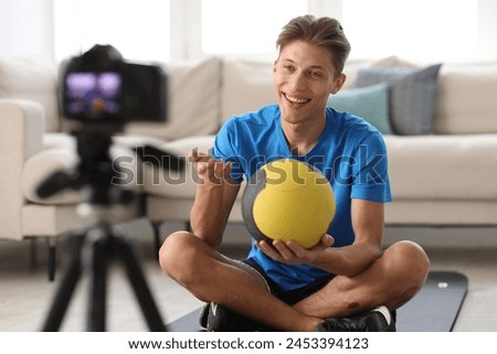 Smiling sports blogger holding medicine ball while recording fitness lesson with camera at home
