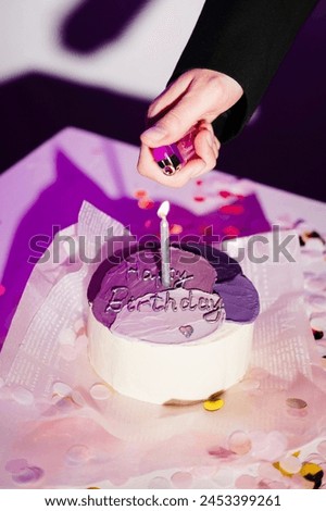 The image shows a hand holding a pink lighter, which is being used to light a candle on a birthday cake. The cake is decorated with purple frosting and has the words "Happy Birthday" written on it. Th