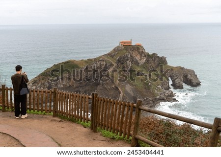 Panoramic view of San Juan de Gaztelugatxe, a small hermitage located in the middle of the sea on the Basque coast, and on the side a Japanese tourist with his back to it taking pictures.