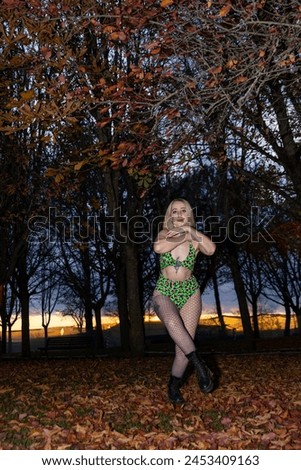 A female dancer in a vibrant neon leopard print bodysuit poses on an autumn leaf-covered ground, with a soft sunset in the background.
