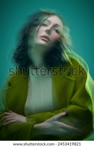 Portrait of a young brunette woman - fashion model posing in motion in a bright olive coat on a green background. Fashion and Art.  Mixed blurry light.