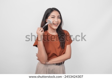 Attractive young Asian woman holding makeup brush to apply blush while getting ready with happy expression isolated white background.