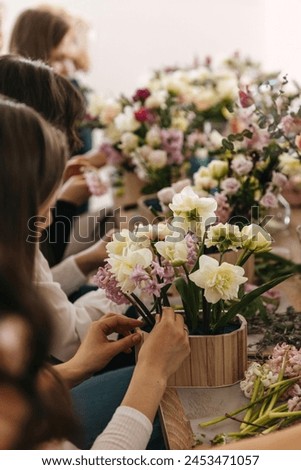 Hands arranging flowers in a floral workshop setting, with a colorful backdrop of various flowers.