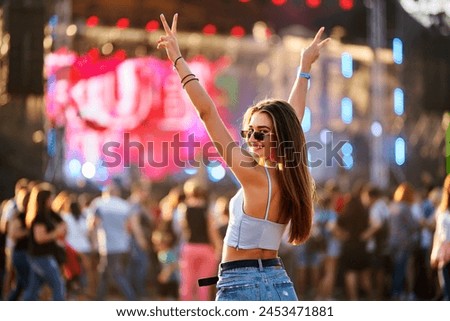 Happy young woman enjoys summer music festival on beach, arms raised, peace sign. Crowd dances near stage in sunlight. Party vibe, live concert enjoyment. Trendy outfit, sunglasses fashion statement.