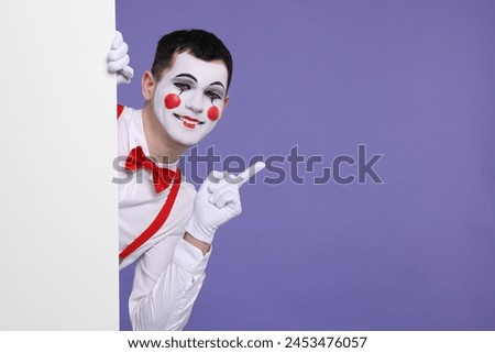 Funny mime artist peeking out of blank poster and pointing at something on purple background. Space for text